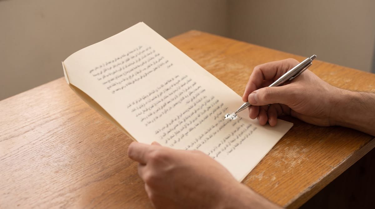 Arabic translation visual: A hand steadying a page flat while the other writes a correction into the margin.